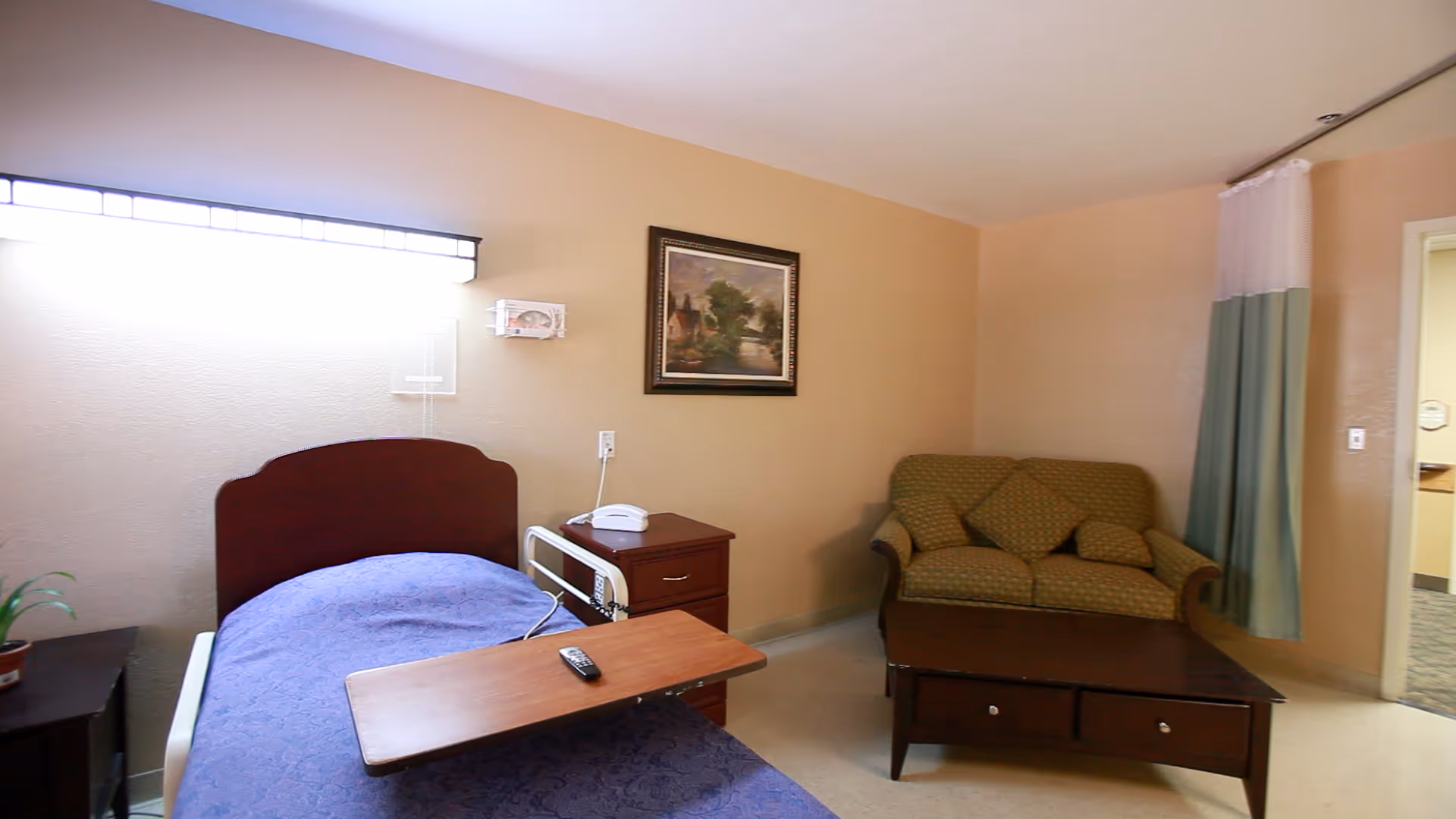 A nursing center room with a single bed covered with a blue blanket, a wooden bedside table with a telephone, a small green couch with two cushions, a wooden coffee table, a framed painting on the wall, and a window with a curtain partially drawn.