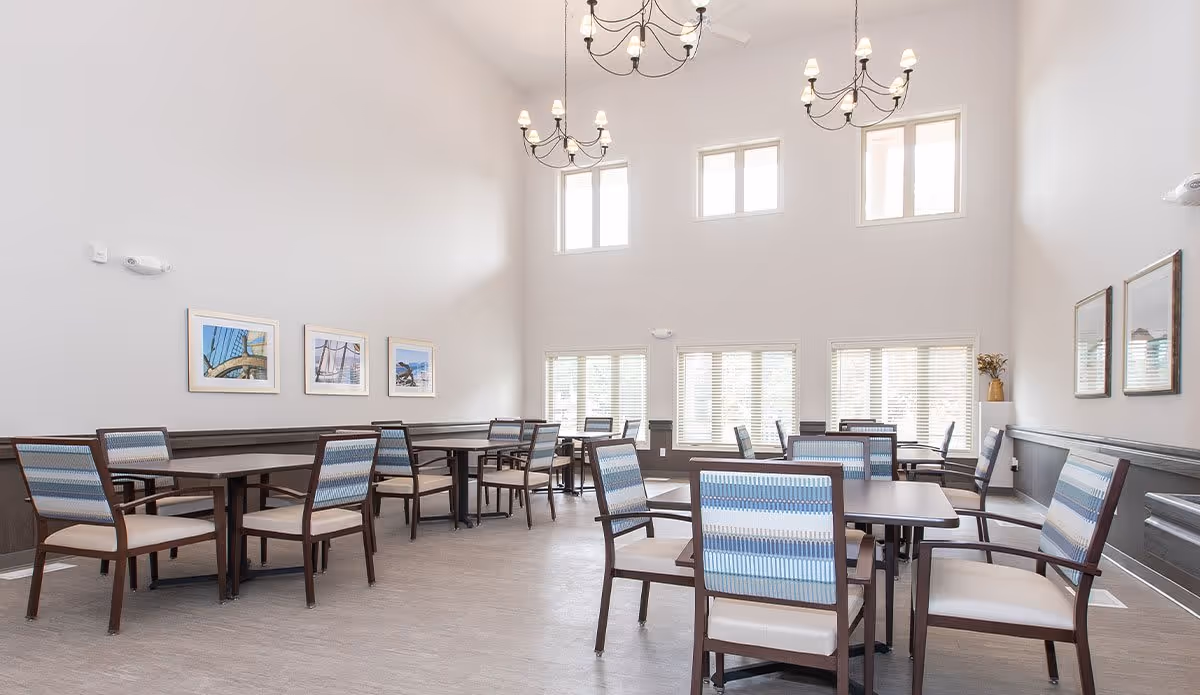 Bright dining room with multiple tables and chairs arranged neatly. The room has high ceilings with three chandeliers, large windows letting in natural light, and framed pictures on the walls.
