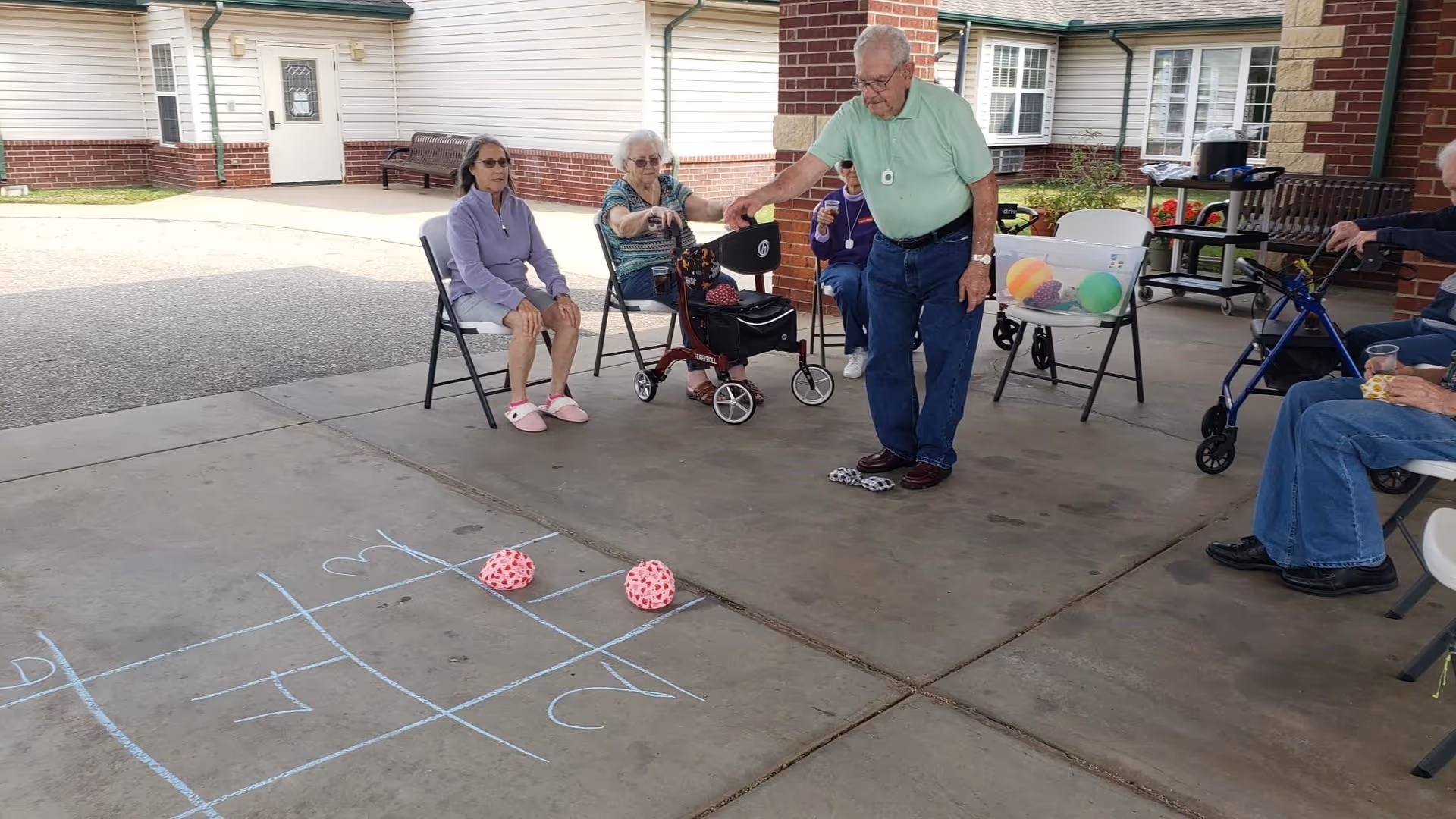 A group of elderly people sitting and standing under a covered outdoor area at a senior living facility. One elderly man is standing and appears to be playing a game involving tossing bean bags onto a chalk-drawn grid on the concrete floor. Other seniors are seated in chairs around him, some with walkers nearby. The building's brick exterior and windows are visible in the background.