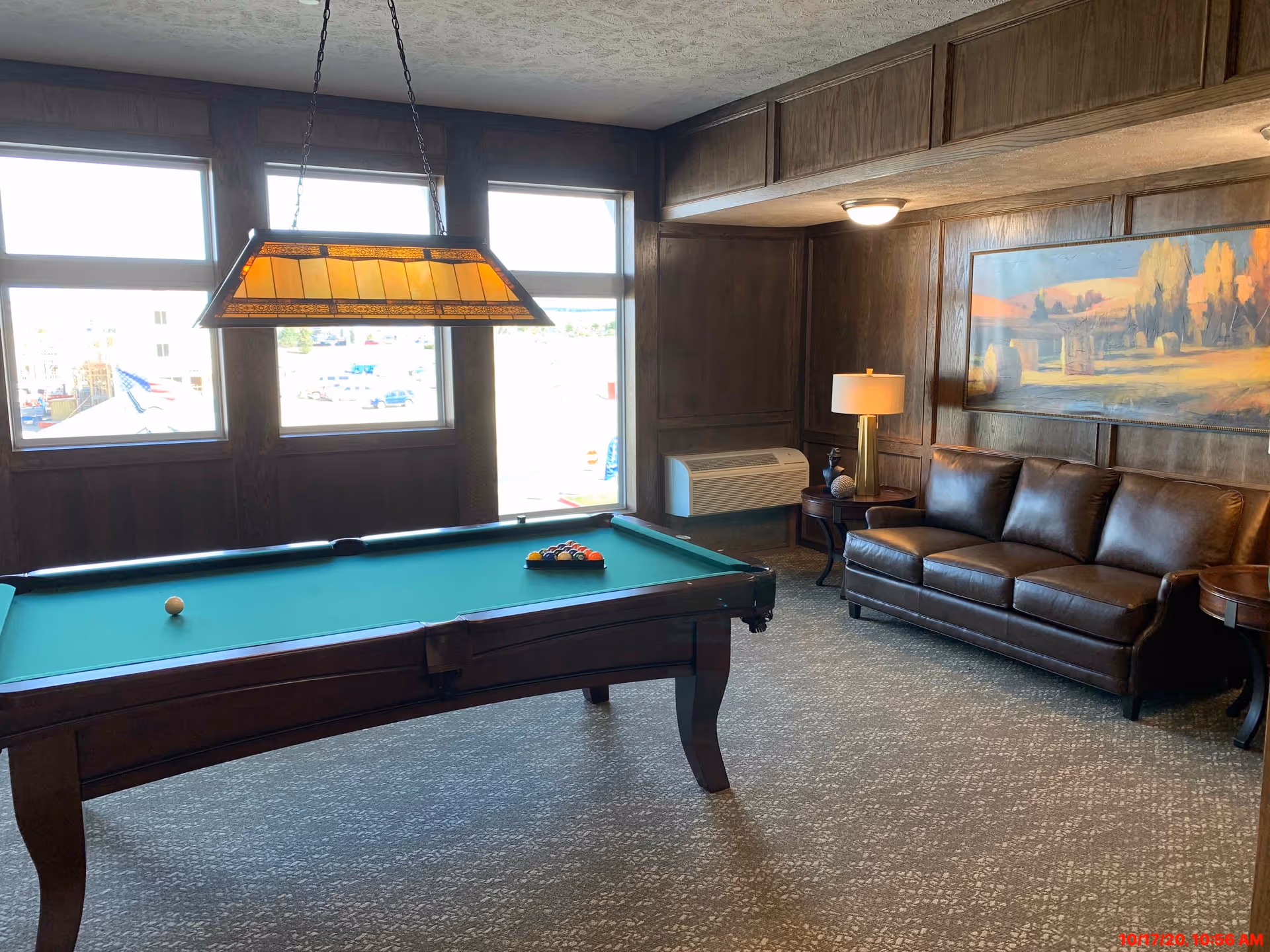 A cozy room with a pool table in the center, a brown leather couch against a wood-paneled wall, a side table with a lamp, and a large landscape painting above the couch. Three large windows let in natural light, and a rectangular hanging light fixture is above the pool table.