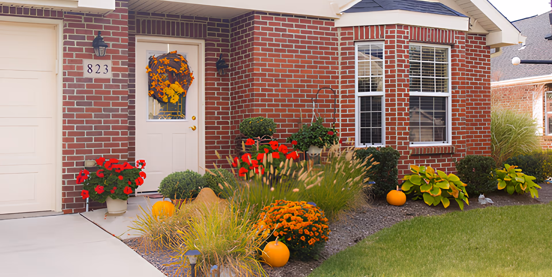 Front entrance of a red brick home with a white door and garage, fall wreath on the door and pumpkins and flowers in the landscaped front yard.
