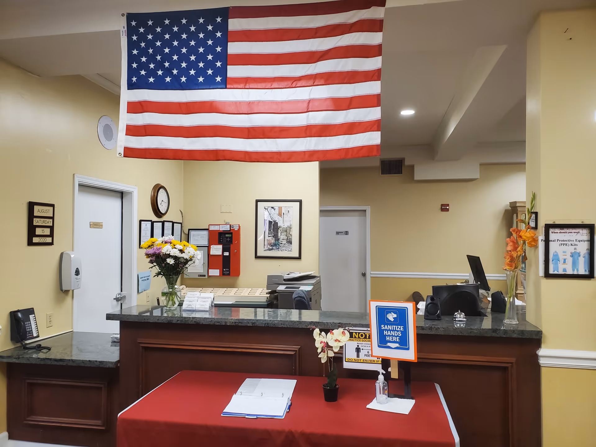 Reception area of a senior living facility with a large American flag hanging from the ceiling. The front desk has a granite countertop with a vase of flowers and a sign instructing to sanitize hands. Behind the desk are office supplies, a clock, a fire alarm panel, and framed notices on the wall. A red table with a guest book and hand sanitizer is in front of the desk. The walls are painted light yellow.