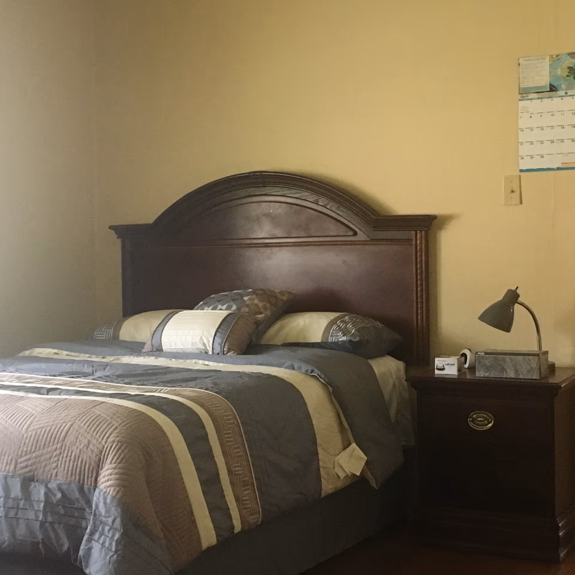 A neatly made bed with a dark wooden headboard, pillows and striped bedding beside a nightstand holding a lamp and small items, with a calendar on the wall.