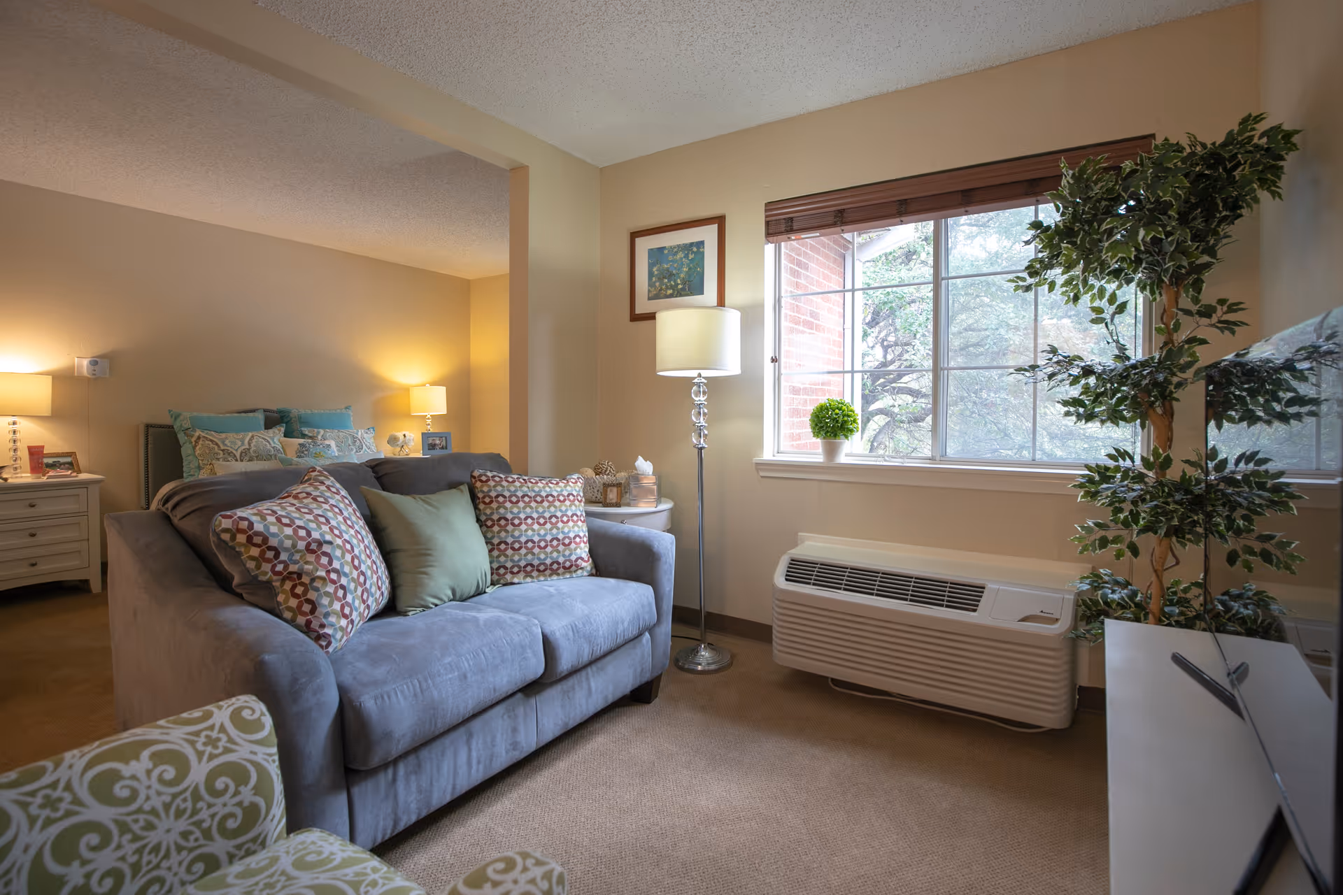 A cozy living room area with a gray couch adorned with patterned and solid pillows, a green patterned armchair, a floor lamp, and a large window with a view of trees outside. In the background, a bedroom with a bed, nightstands, and lamps is visible.