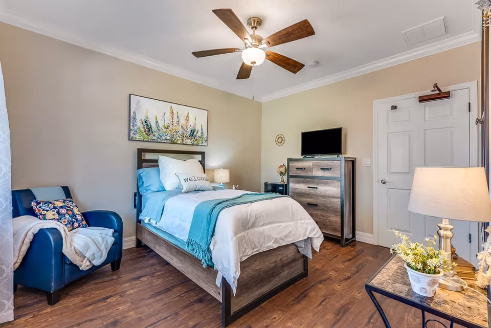 A cozy senior living bedroom with a single bed dressed in white and teal bedding, a 'welcome' pillow, a blue armchair with a floral cushion and blanket, a wooden dresser with a TV on top, a side table with a lamp and a potted plant, and a ceiling fan with light. The room has wooden flooring and beige walls with a floral painting above the bed.