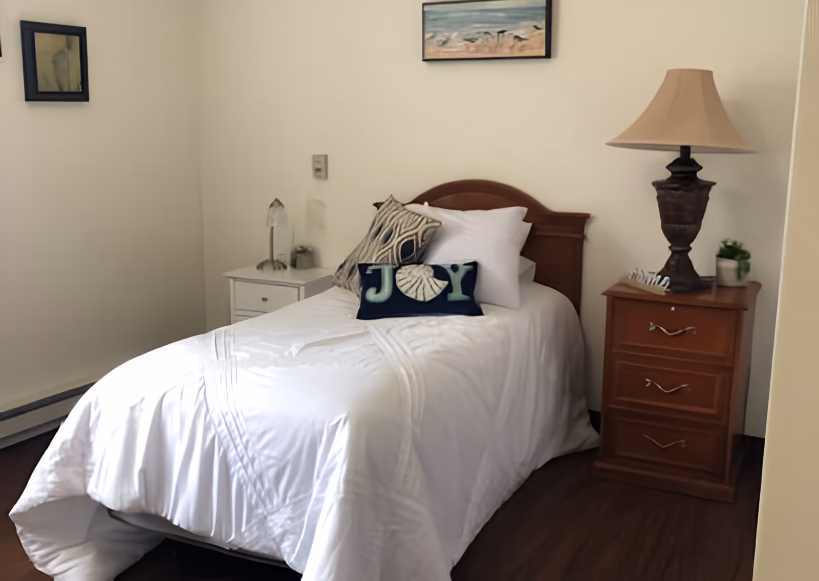 A neatly made single bed with white bedding and decorative pillows, including one with the word 'JOY'. The bed has a wooden headboard and is flanked by two nightstands. The left nightstand is white with a small lamp and decorative items, while the right nightstand is wooden with three drawers, a lamp, and a small plant. A framed picture hangs on the wall above the bed.