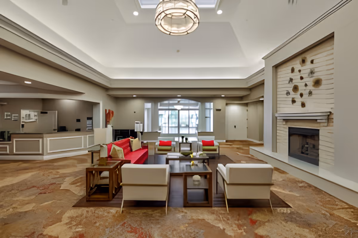 A spacious and well-lit living room area in a senior living facility featuring a red sofa, two white armchairs, two green chairs, a wooden coffee table, and a fireplace with decorative art above it. The room has a high ceiling with recessed lighting and a large window in the background letting in natural light.