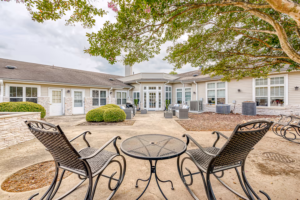 Outdoor courtyard of a senior living facility with a metal patio table and chairs facing the building entrance and seating areas.