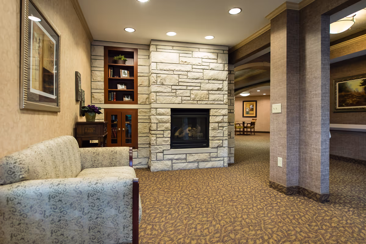 A cozy interior common area with a patterned sofa on the left, a stone fireplace in the center, built-in wooden shelves with books and plants, and a carpeted floor. The area is softly lit with recessed ceiling lights and has framed artwork on the walls.