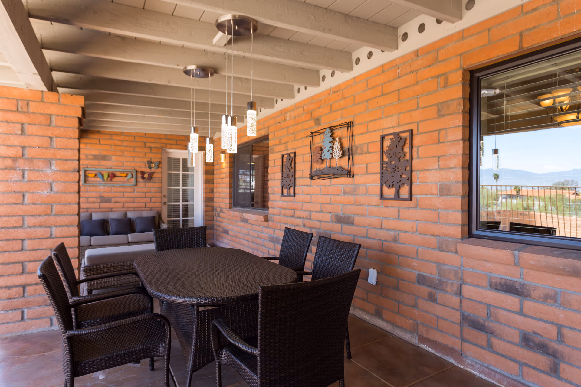 Covered outdoor patio with a wicker dining table and chairs, sofa seating, hanging pendant lights, and brick walls with decorative wall art.