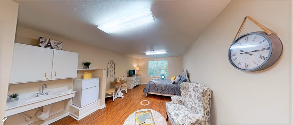 Interior view of a studio apartment with a bed, armchair, kitchenette with sink and mini fridge, dresser and wall clock.