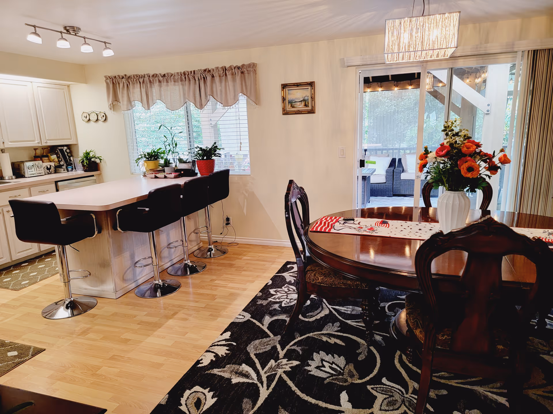 A bright and cozy dining and kitchen area featuring a wooden dining table with a floral centerpiece, surrounded by ornate chairs on a patterned rug. The kitchen has a large island with four black bar stools, white cabinets, and various potted plants on the windowsill. A sliding glass door leads to an outdoor patio with seating visible outside.
