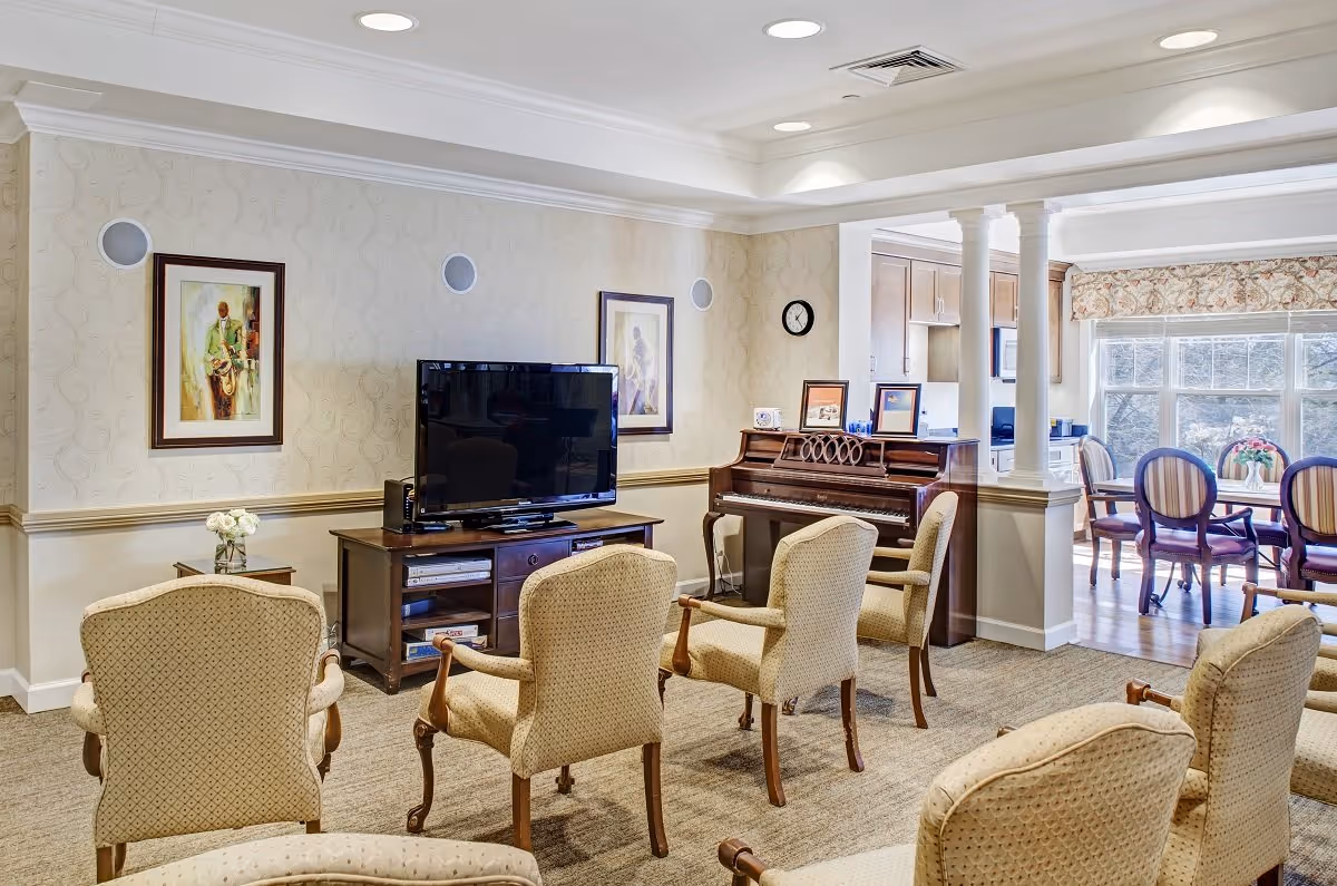Bright senior living common room with upholstered chairs facing a TV, a piano against the wall, and a dining area visible through columns.