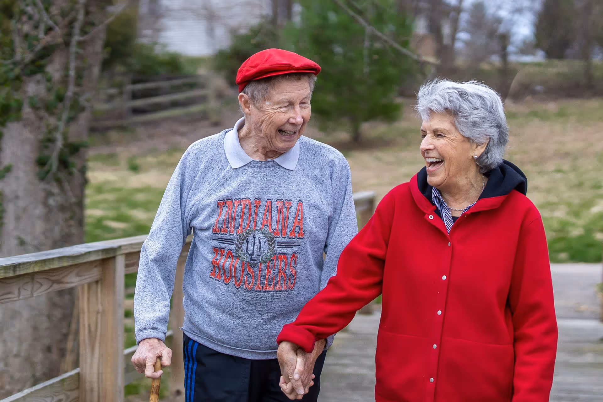 An elderly man and woman walking outdoors on a wooden path, holding hands and smiling at each other. The man is wearing a red cap and a gray Indiana Hoosiers sweatshirt, while the woman is wearing a red jacket. Trees and grass are visible in the background.