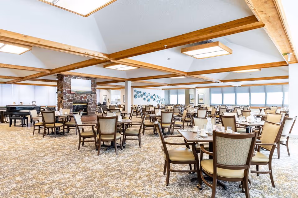 Spacious dining room with multiple wooden tables and chairs arranged neatly. The room features a stone fireplace with a painting above it, large windows with blinds, and wooden beams on the ceiling. The carpet has a patterned design, and the room is well-lit with ceiling lights.