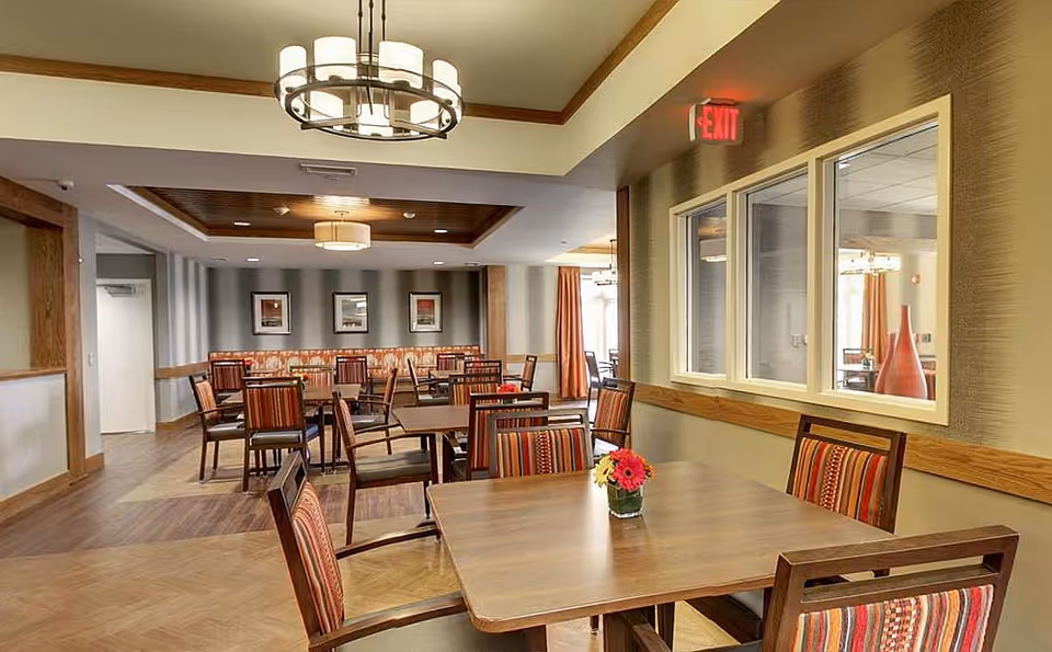 A well-lit dining room in Evergreen Senior Living with multiple wooden tables and chairs featuring striped upholstery. The room has wooden flooring, framed artwork on the far wall, large windows on the right, and modern ceiling light fixtures. A small vase with colorful flowers is placed on the nearest table.