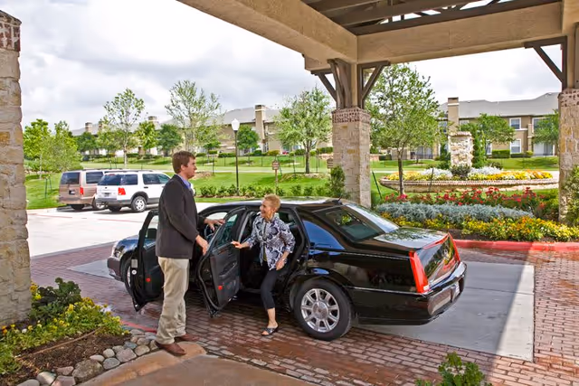 A man in a suit is helping an elderly woman step out of a black car under a covered entrance with stone pillars. In the background, there is a landscaped garden with flowers, trees, and a fountain, along with a large residential building.