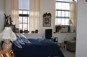 A cozy bedroom with a blue bedspread, two large windows with cream-colored curtains, a small TV on a stand, a framed picture on the wall, and various decorative items including a lamp and plants.