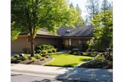 A single-story residential building with a sloped roof surrounded by lush green trees and well-maintained landscaping. There is a green lawn, shrubs, and a paved walkway leading to the entrance of the building.