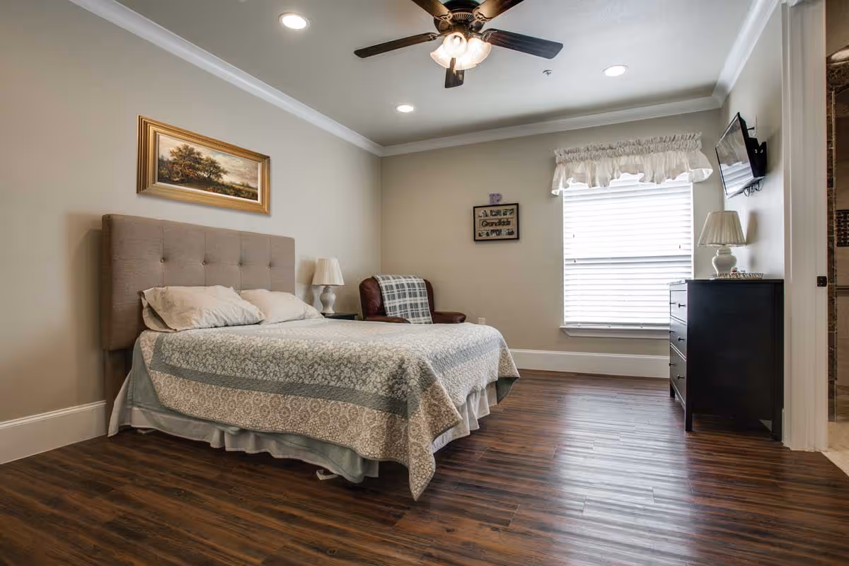 Bright bedroom with a tufted headboard bed, nightstand and lamp, armchair, dresser with a wall-mounted TV, ceiling fan, and wood floors.