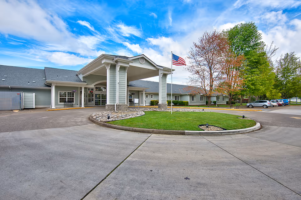 Front exterior view of a single-story assisted living facility with a covered entrance, an American flag on a flagpole in a circular grassy area, several trees with green and autumn-colored leaves, and a parking area with cars under a partly cloudy blue sky.