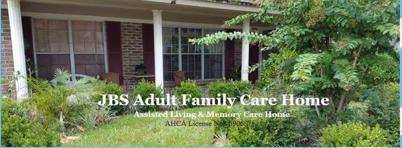 Front exterior view of JBS Adult Family Care Home showing a porch with white columns, large windows, and surrounding green bushes and plants.