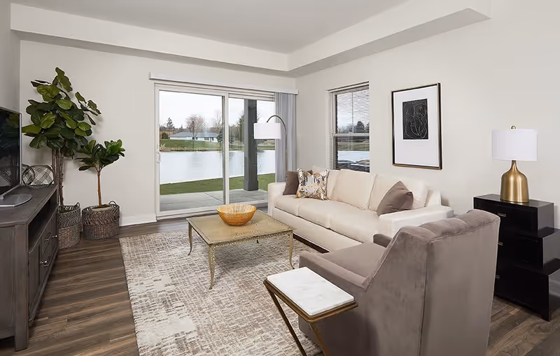 Bright living room with a sofa, armchair, coffee table and TV console facing a sliding glass door overlooking a pond.