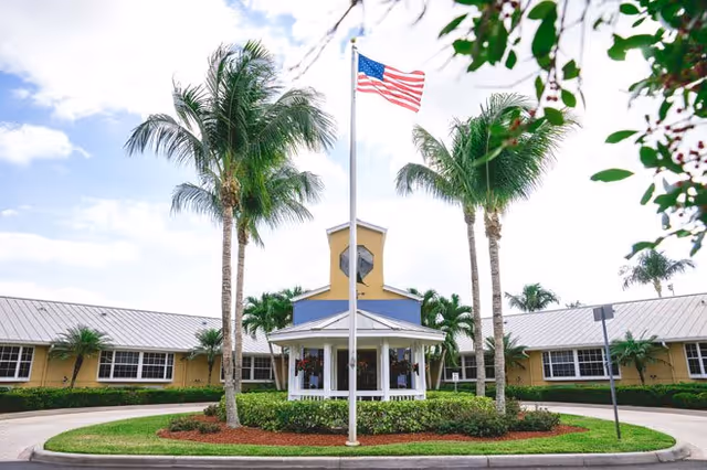 Exterior view of Barrington Terrace of Fort Myers facility featuring a circular driveway with a landscaped center island containing palm trees, a white gazebo, and an American flag on a flagpole. The building is single-story with a light yellow facade and a metal roof under a partly cloudy sky.