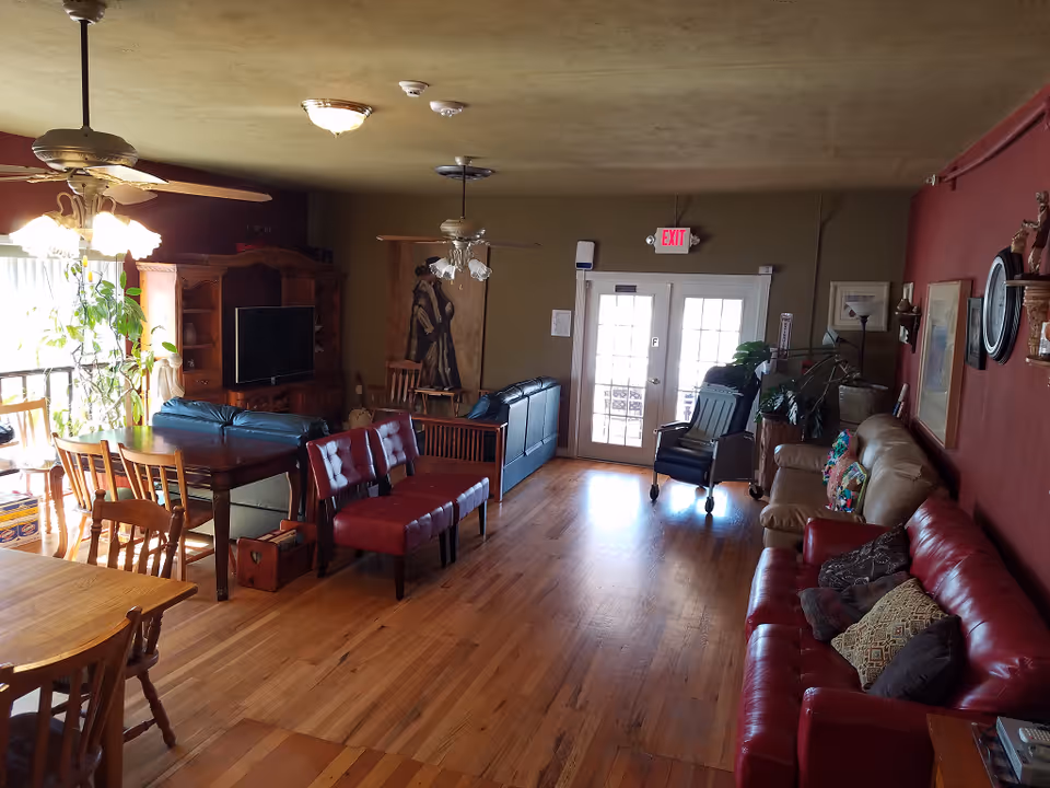 A spacious living room in an assisted living home with wooden floors, multiple seating options including red and beige sofas, a black recliner, and wooden chairs around a dining table. The room has ceiling fans with lights, a large wooden entertainment center with a TV, plants near a window, and French doors leading outside. The walls are decorated with paintings and a clock.