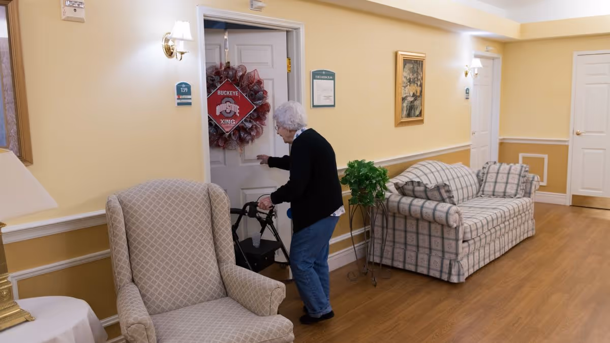 An older adult using a walker opens a door in a furnished senior living hallway with an armchair and sofa.