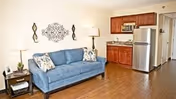 A cozy living room area with a blue sofa adorned with patterned pillows, two side tables each with a lamp, decorative wall art above the sofa, and a kitchen area in the background featuring wooden cabinets, a microwave, and a stainless steel refrigerator.