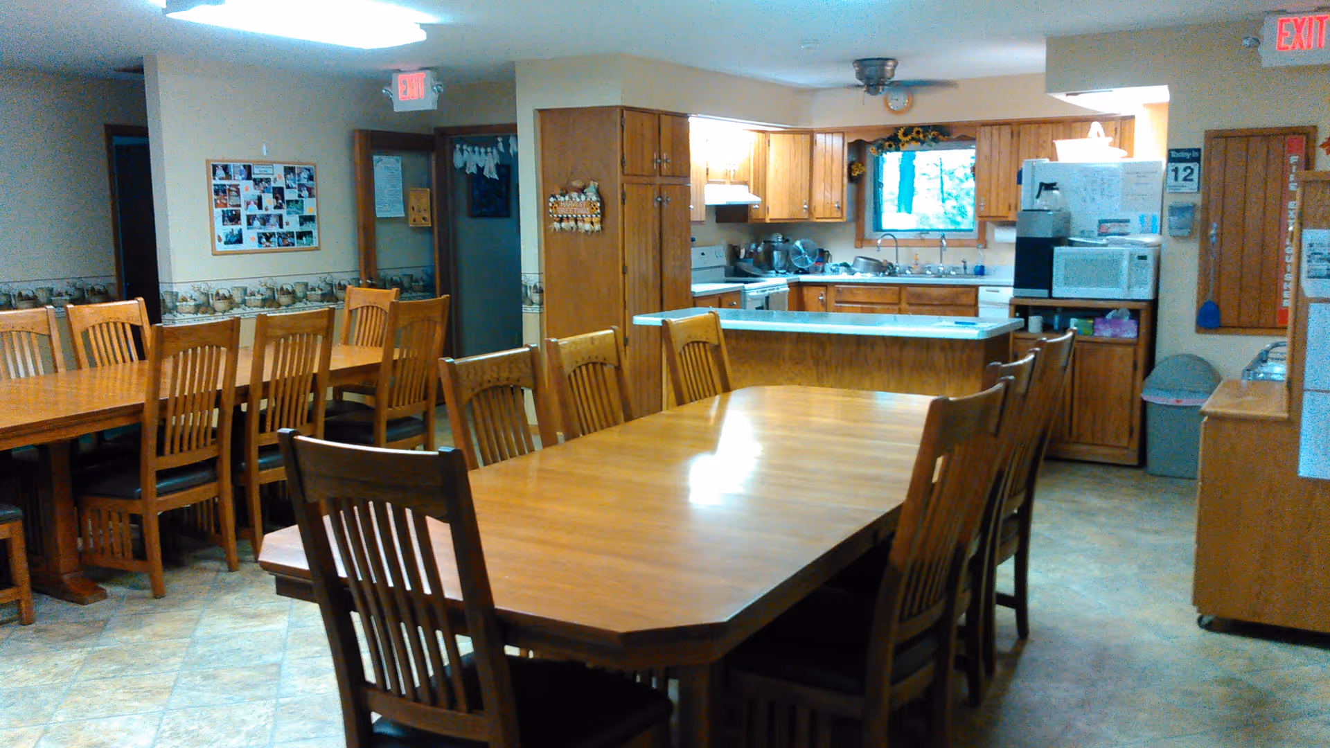 Interior view of a senior living facility dining area with two large wooden tables surrounded by wooden chairs. In the background, there is a kitchen area with wooden cabinets, a stove, a microwave, a refrigerator, and a window with a sunflower-themed valance. The floor is tiled, and there are bulletin boards and an exit sign visible on the walls.