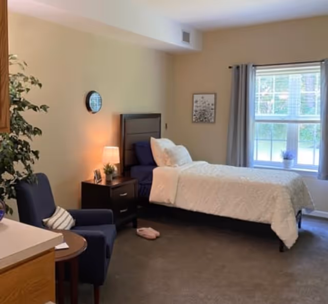 Well-lit senior living bedroom with a twin bed, nightstand and lamp, an armchair, and a large window.
