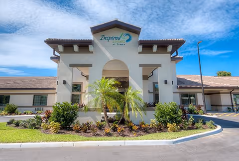 Front exterior view of the Inspired Living Tampa senior living facility building with a landscaped garden featuring palm trees and shrubs under a blue sky with some clouds.