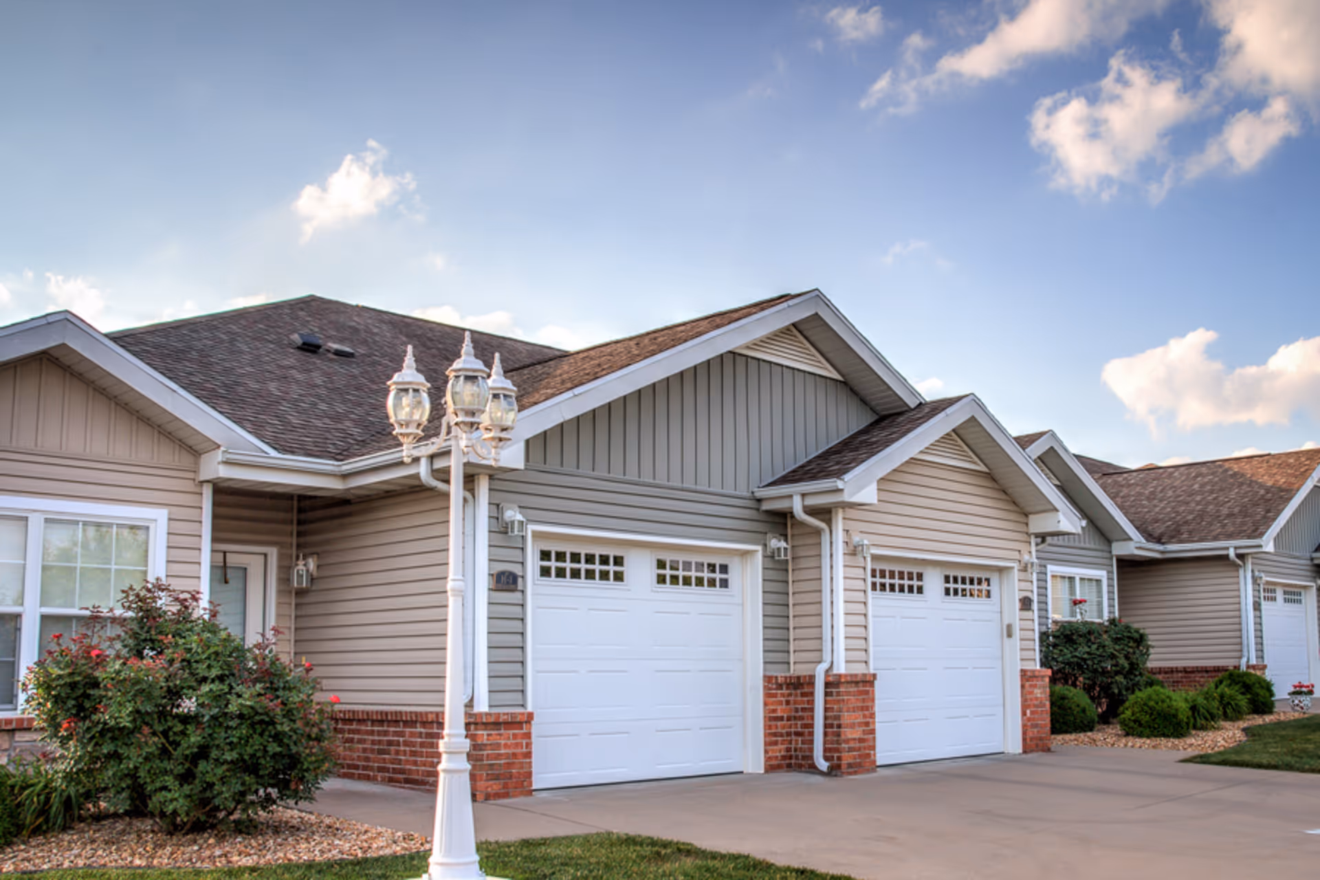 Exterior view of a senior living facility showing a row of attached single-story units with beige siding, brick accents, white garage doors, and a decorative white street lamp in the foreground under a partly cloudy sky.
