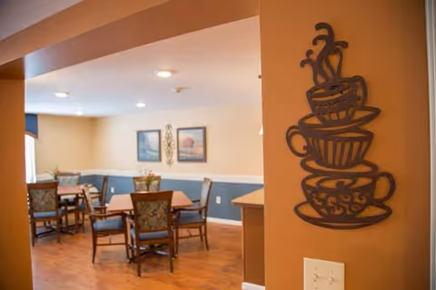 Interior view of a dining area in a senior living facility with wooden tables and chairs, decorative wall art, and a metal wall decoration shaped like stacked coffee cups on an orange wall.