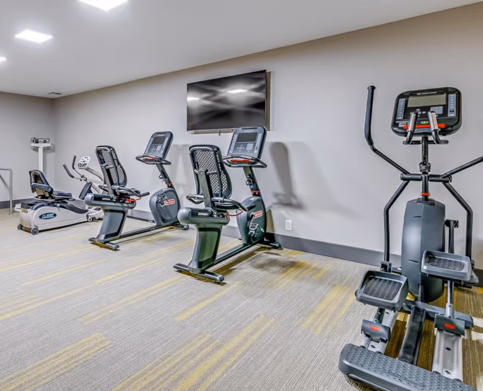 Small fitness room with several exercise bikes and an elliptical facing a wall-mounted TV.