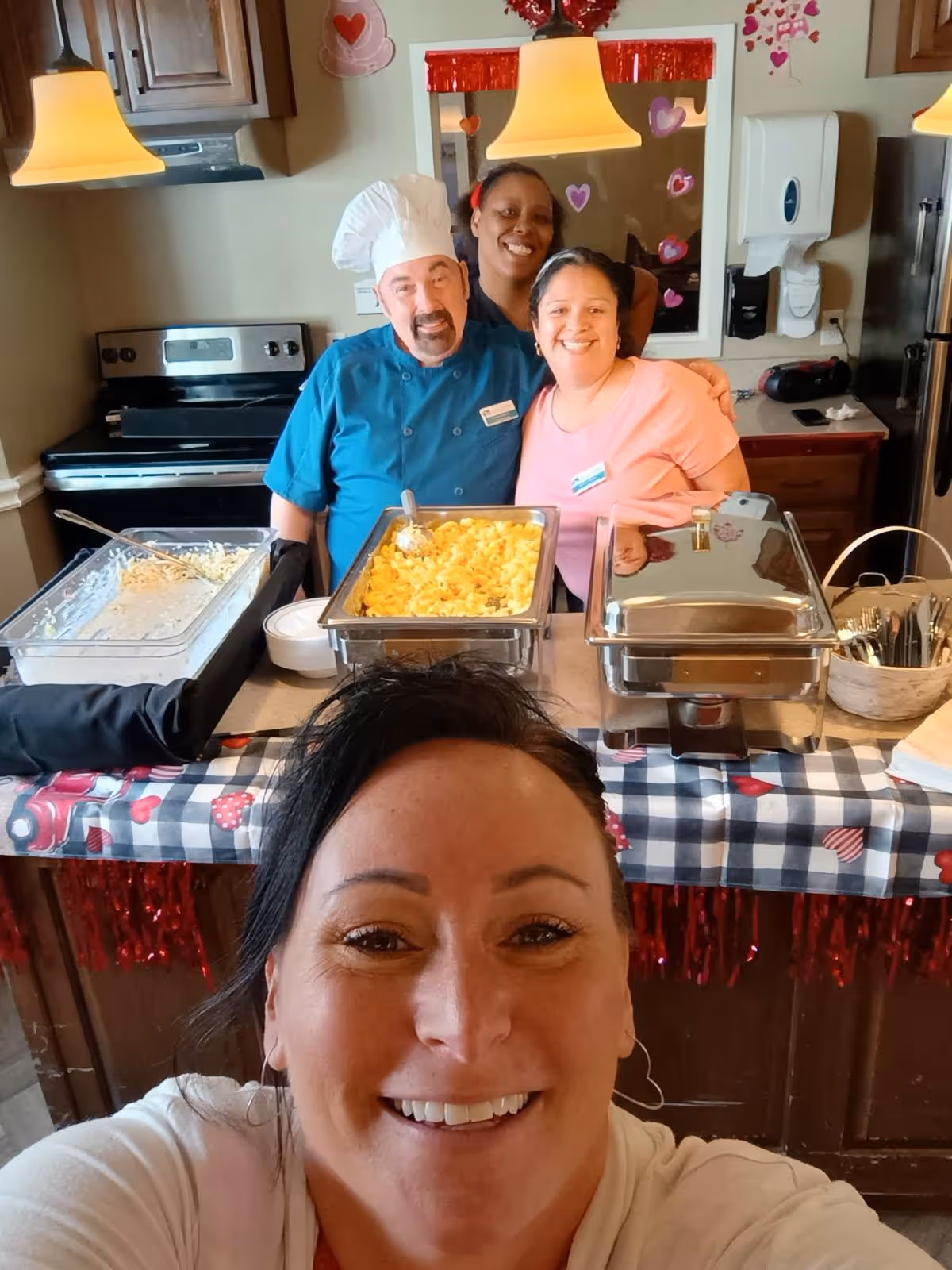 A group of four smiling people posing for a selfie in a kitchen area. Three people stand behind a counter with trays of food, including macaroni and cheese, while the person taking the selfie is in the foreground. The kitchen has wooden cabinets, a stove, and a refrigerator. The background is decorated with red and pink heart decorations.