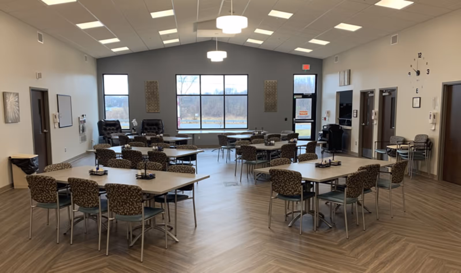 Spacious multipurpose common room with several tables and patterned chairs arranged for dining or activities, a large window and seating area at the far wall.
