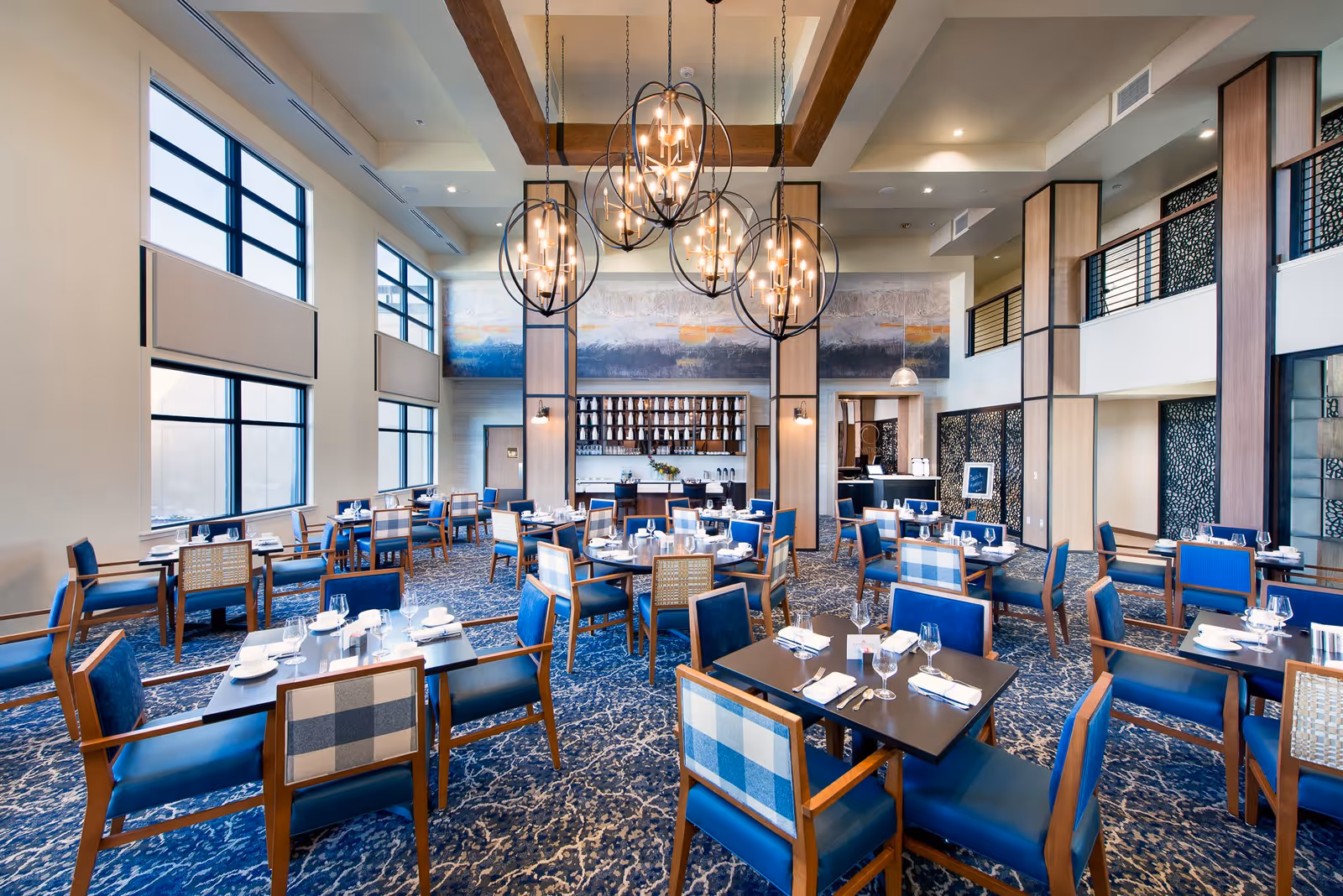 A spacious and elegant dining room with multiple tables set with white plates, glasses, and silverware. The room features large windows allowing natural light, blue cushioned chairs with wooden frames, patterned carpet, and modern spherical chandeliers hanging from a high ceiling with wooden beams. The back wall has a large abstract painting and a shelf with decorative items.
