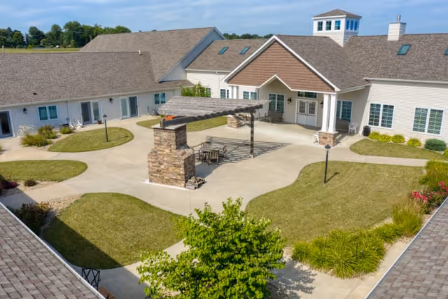 Outdoor courtyard area of a senior living facility with a pergola-covered seating area, stone pillars, concrete walkways, green lawns, and surrounding buildings under a blue sky.