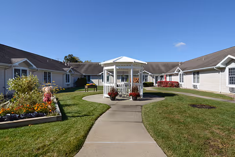 Outdoor courtyard area of a senior living facility with a central white gazebo surrounded by a concrete walkway and green grass. The courtyard is bordered by single-story beige buildings with white trim under a clear blue sky. There are flower beds with colorful plants and a scarecrow decoration on the left side.