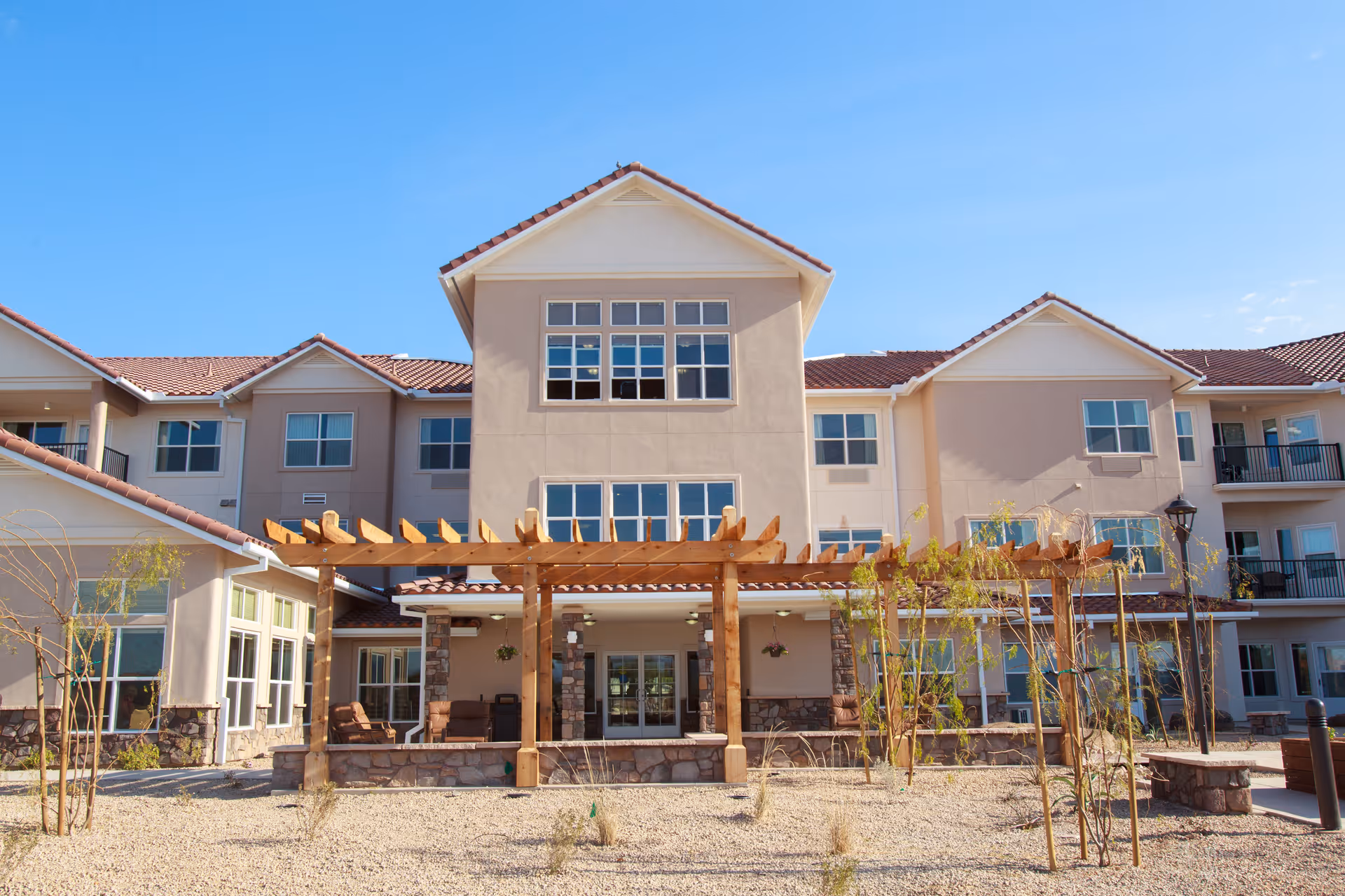 Exterior view of a multi-story senior living facility with beige walls and red-tiled roofs under a clear blue sky. The building features multiple windows and balconies, with a wooden pergola structure and some young trees and shrubs in the foreground.