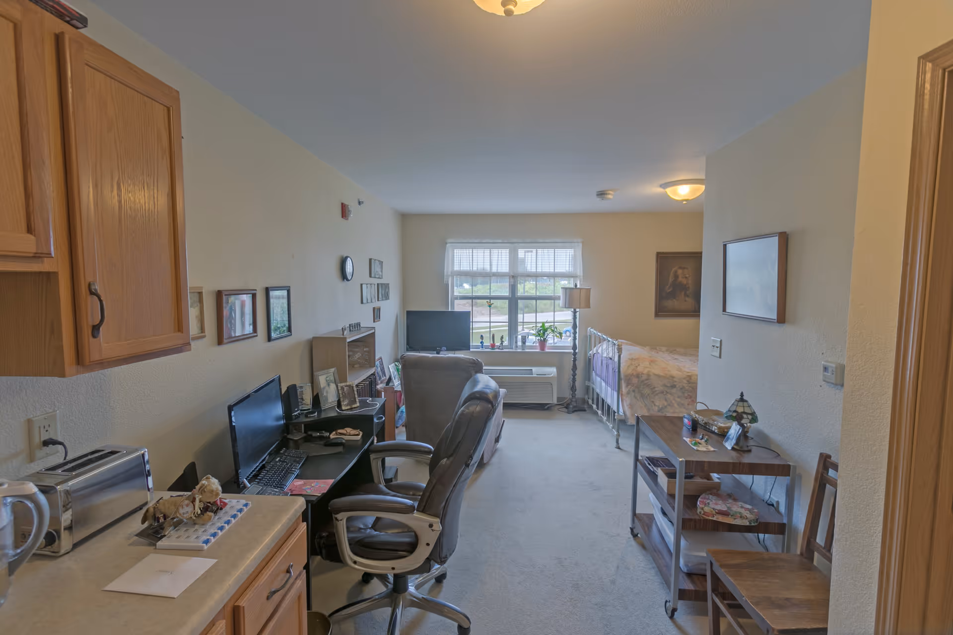 Interior view of a senior living facility room at Heritage Woods of Watseka, showing a combined living and sleeping area. The room includes a bed near a window with blinds, a TV on a stand, a desk with a computer and office chair, wooden cabinets, and various framed pictures on the walls. There is also a small table with chairs and a lamp, creating a cozy and functional living space.