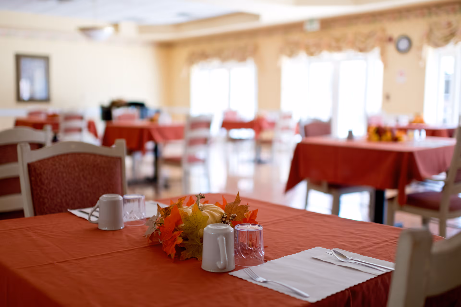 A dining room with tables covered in red tablecloths, set with white mugs, glasses, and silverware on white napkins. The tables have autumn-themed centerpieces with leaves and small pumpkins. The room is softly lit with large windows in the background letting in natural light.