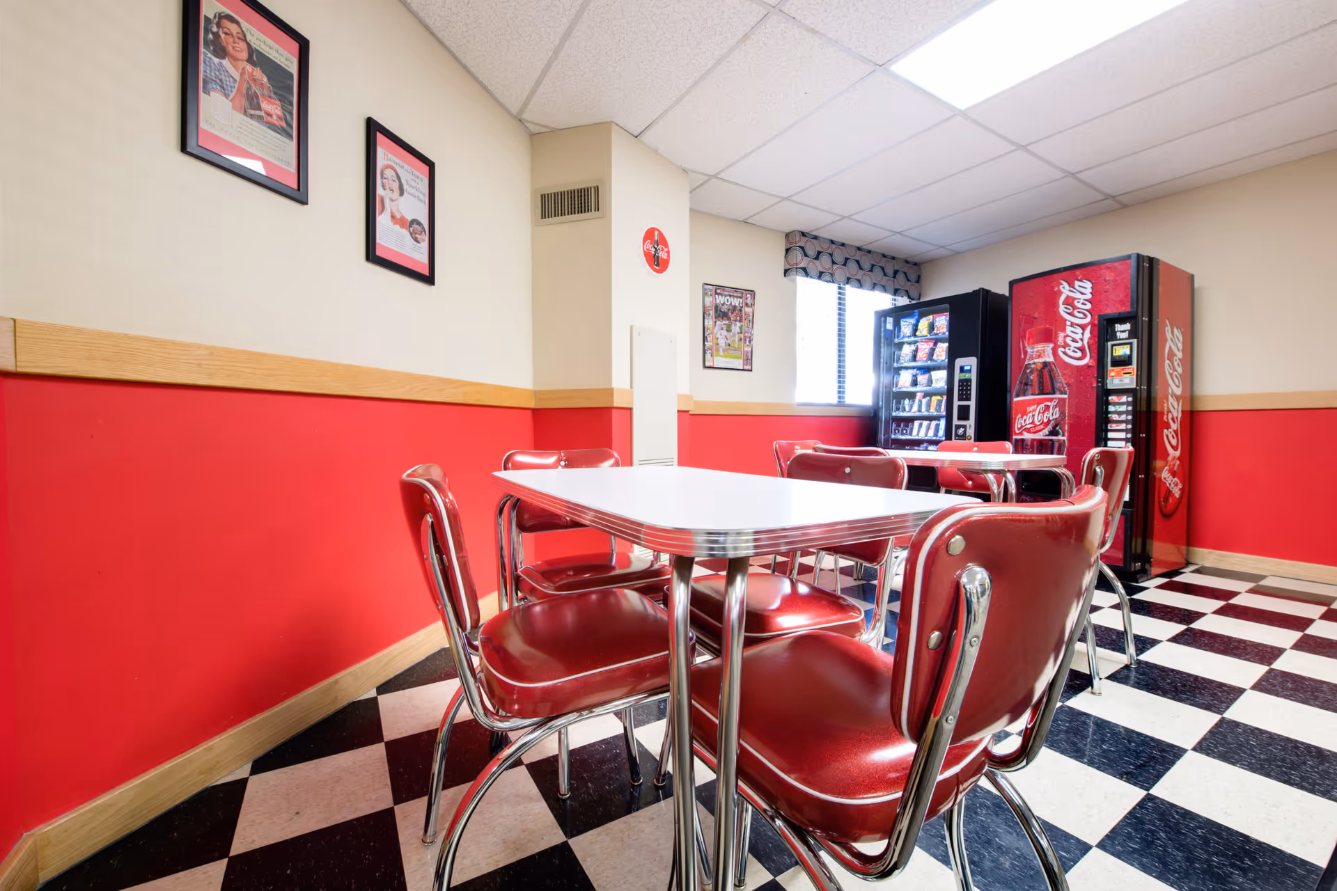 Retro-style break room with red diner chairs around a white table, checkered black-and-white floor, and vending machines against red and beige walls.