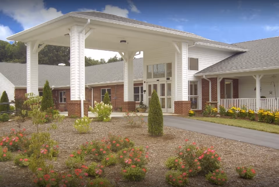 Front entrance of a single-story building with a covered porte-cochere, brick and white siding, and landscaped flower beds.