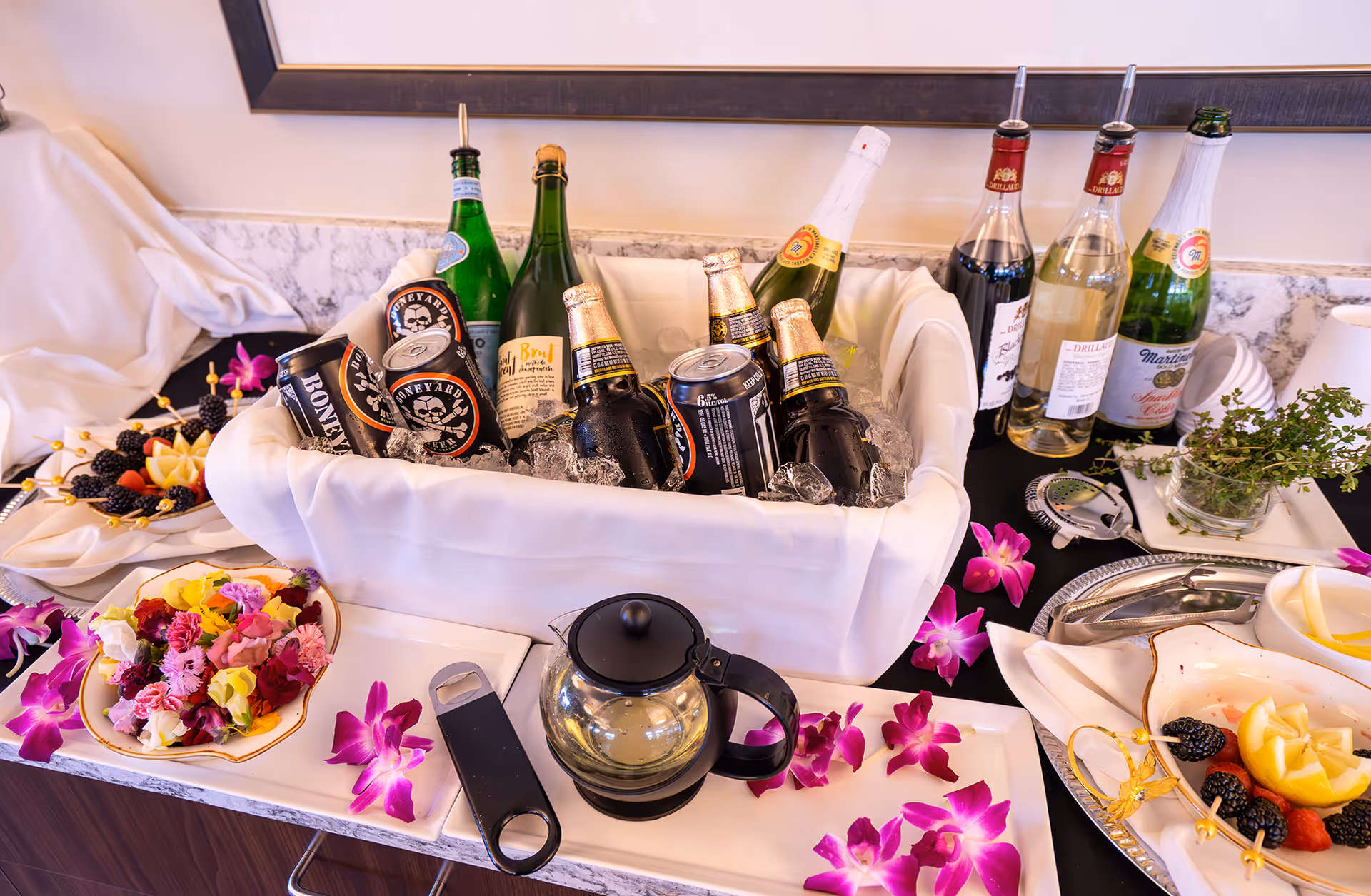 A beverage station with a white cloth-lined ice bucket containing bottles and cans of beer and sparkling water. Surrounding the bucket are plates with colorful flower petals and fruit garnishes, a glass teapot, a bottle opener, and additional bottles of wine and sparkling beverages on a marble countertop. Purple orchid flowers are scattered decoratively around the setup.