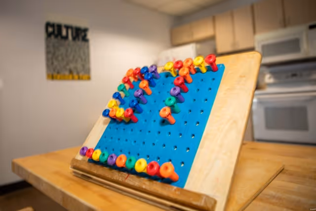 A close-up of a wooden pegboard with colorful pegs arranged on a blue felt surface, placed on a wooden table in a room with a kitchen area in the background including cabinets, a microwave, and an oven. A blurred poster with the word 'CULTURE' is visible on the wall.