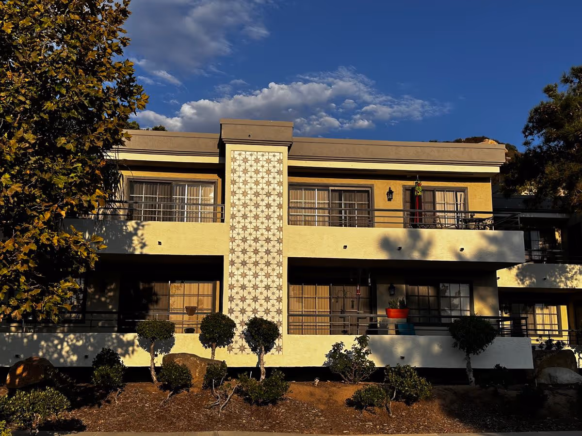 Two-story residential building exterior with balconies, a decorative tiled center panel, and landscaping under a blue sky.